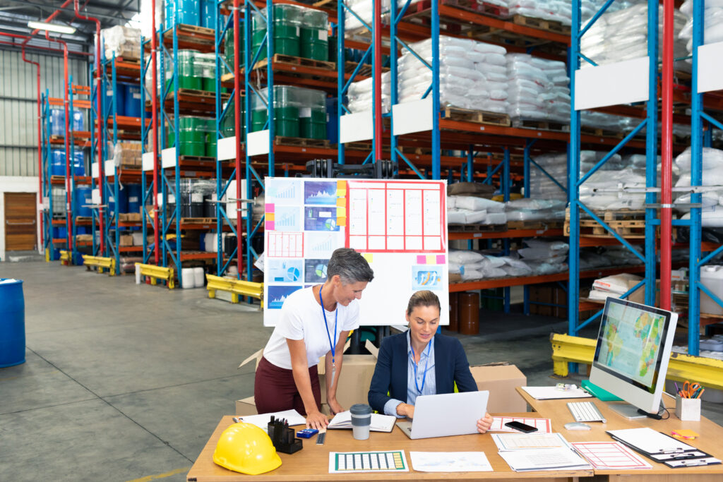 high angle view of beautiful caucasian female manager with her coworker discussing over laptop at desk in warehouse. this is a freight transportation and distribution warehouse. industrial and industrial workers concept