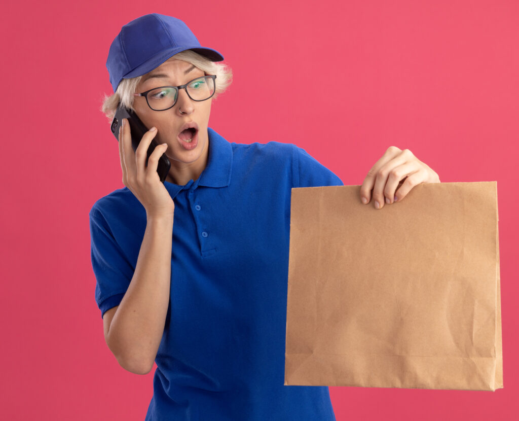 young delivery woman in blue uniform and cap holding paper package looking surprised and amazed while talking on mobile phone over pink background